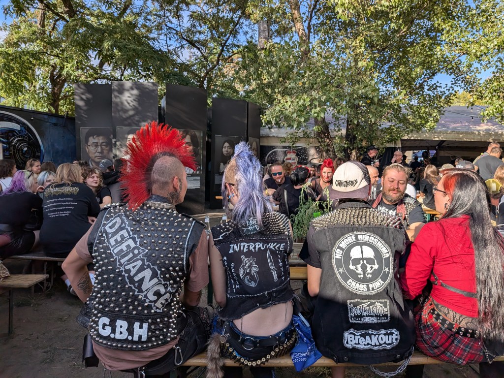 A group of people with punk hairstyles and vests sit at a wooden table in a lively outdoor gathering, surrounded by trees and art displays.
