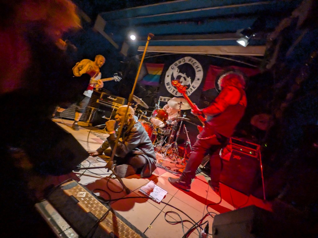 A punk rock band performing on stage with energetic musicians, featuring a vocalist in a kneeling position and a guitarist playing passionately, surrounded by instruments and amplifiers in a dimly lit venue.
