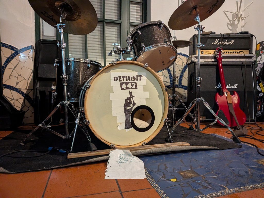 A drum set featuring a bass drum with a 'Detroit 442' sticker, placed next to a guitar and amplifiers in a music venue.